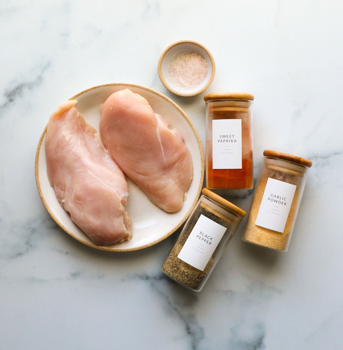 raw chicken breasts on a plate with spice jars of salt, paprika, garlic powder, and black pepper.