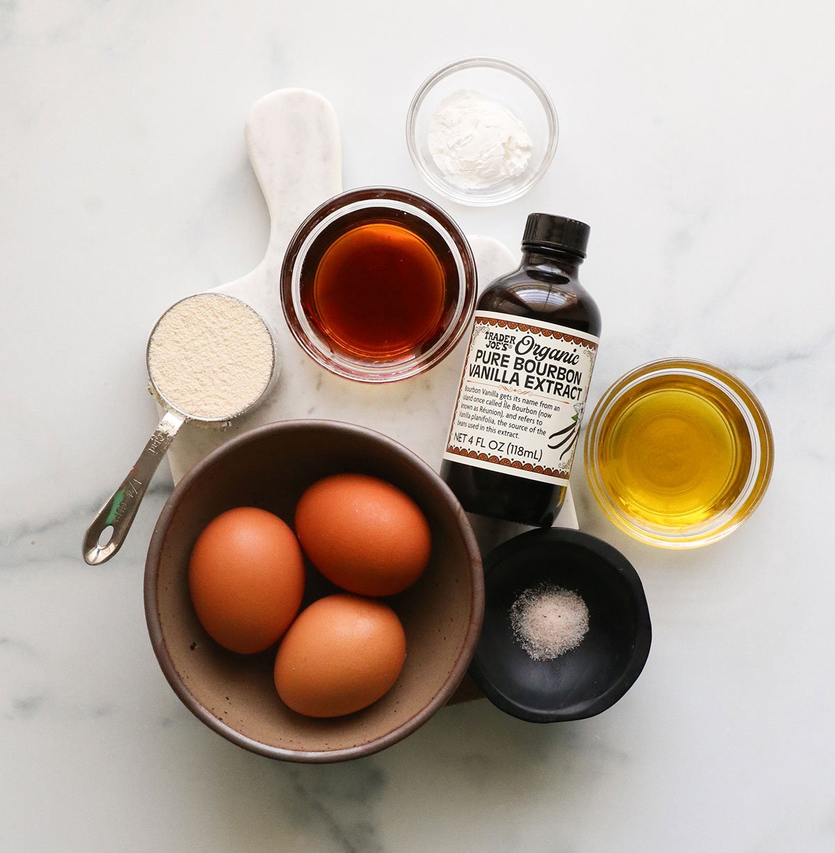 eggs, coconut flour, maple syrup, vanilla, olive oil and salt on a white board.