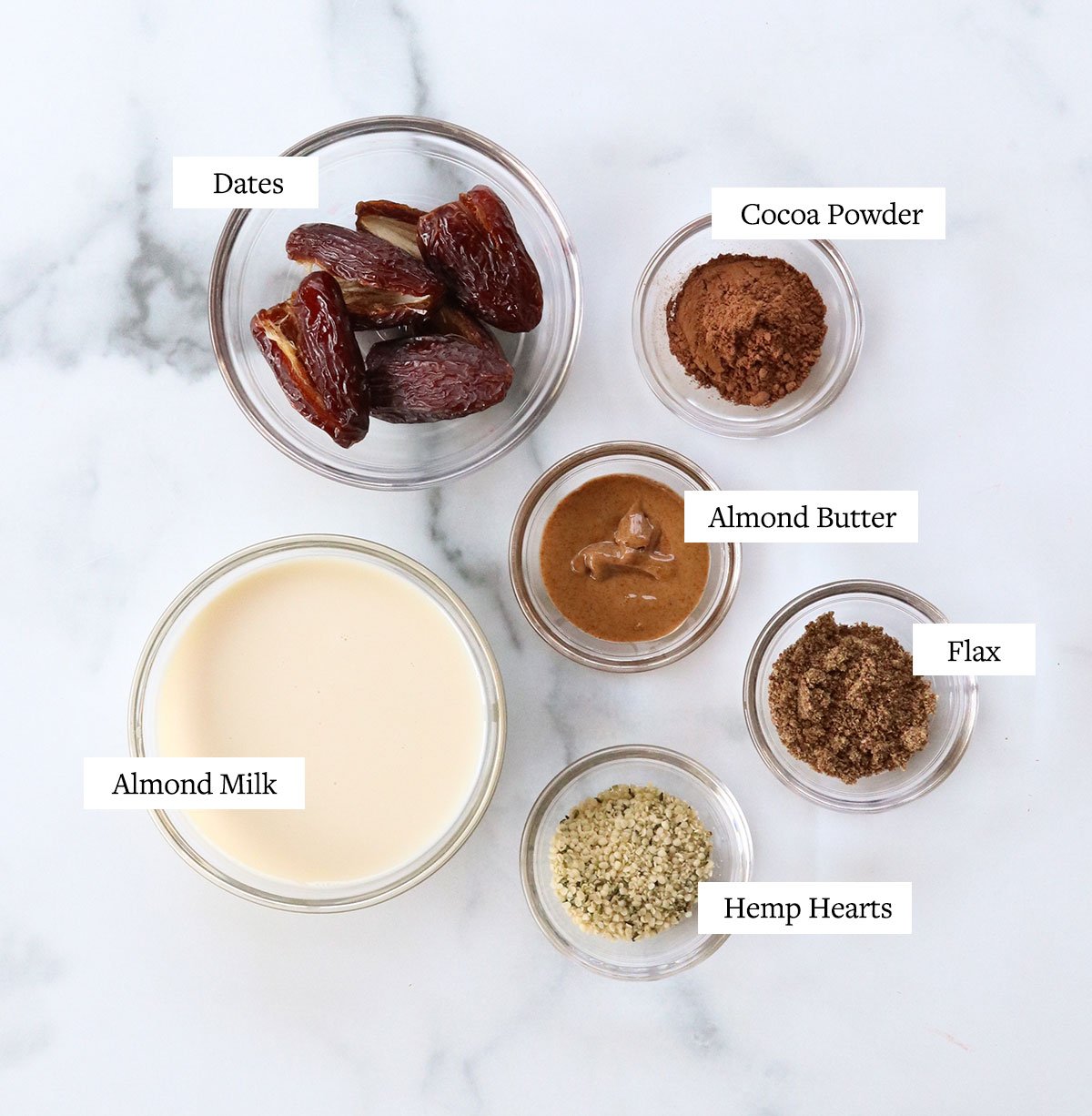 date smoothie ingredients labeled in glass bowls on a white surface.