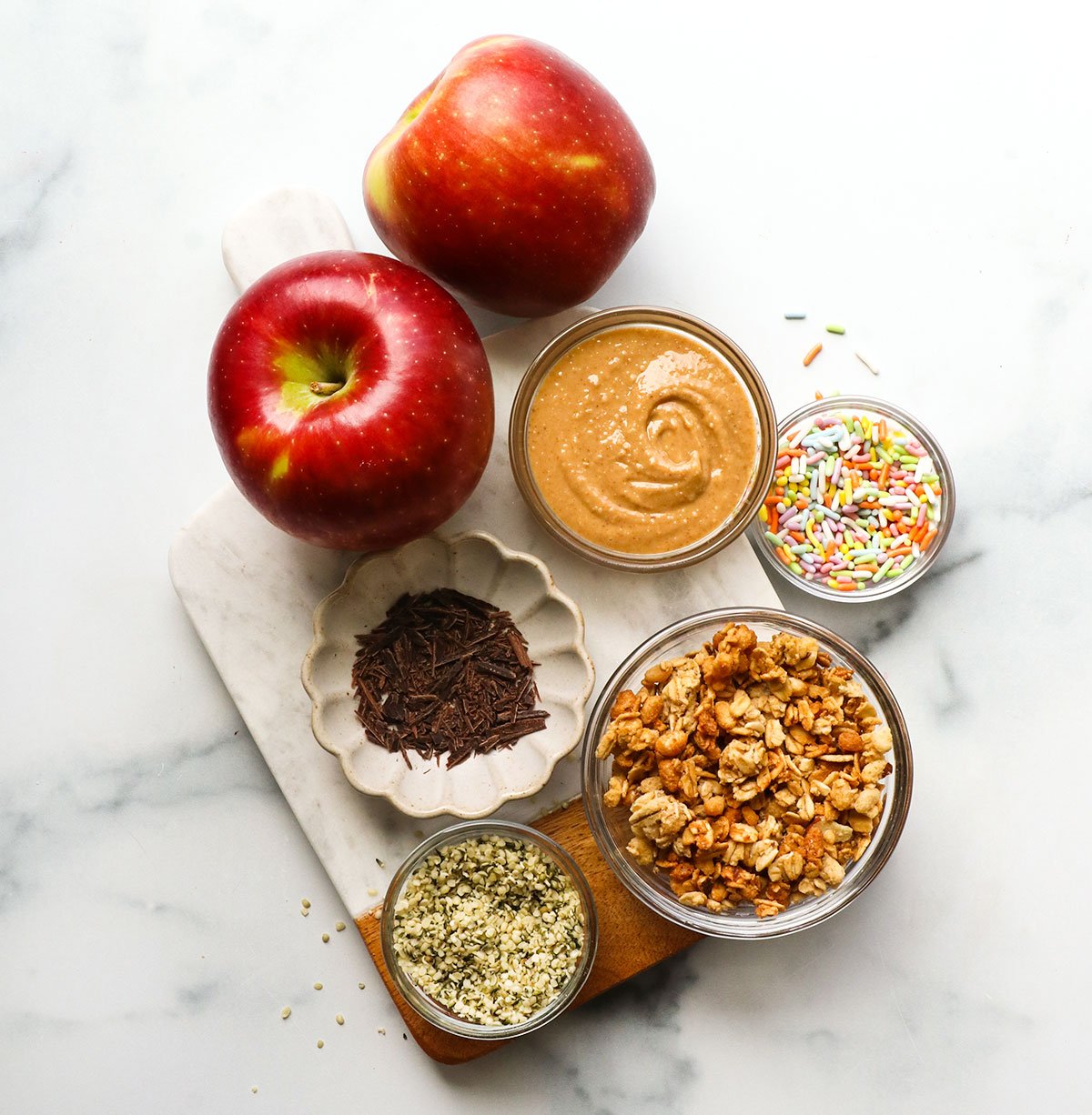 two apples on a board near sprinkles, peanut butter, granola, and hemp hearts.