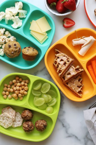 healthy snacks like waffles, fruit, carrots, and turkey on colorful divided plates.