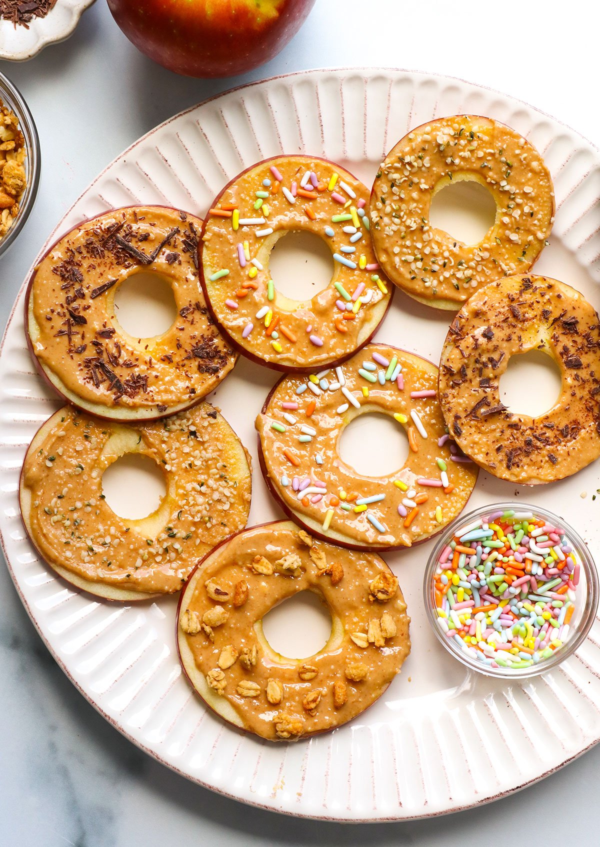 apple slices that look like donuts with sprinkles, granola, hemp hearts, and shaved chocolate on top.