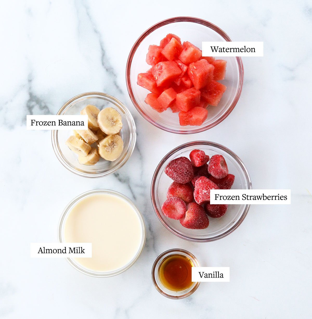 watermelon smoothie ingredients labeled in glass bowls on a white surface.